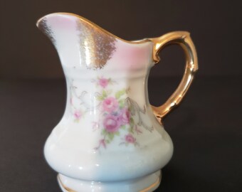 May include: A small white porcelain creamer with a pink and gold floral design. The creamer has a gold handle and a gold rim.