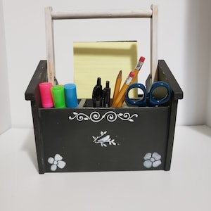 Puede incluir: Un organizador de escritorio de madera negro con un asa blanca. Tiene un diseño floral blanco y está lleno de lápices, bolígrafos y rotuladores.