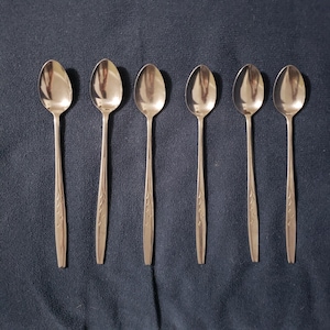 May include: Six silver teaspoons with ornate designs on the handles. The spoons are arranged in a row on a dark blue surface.