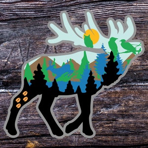 May include: A sticker depicting an elk silhouette with a forest scene inside. The elk is black with a grey outline. The forest scene includes green and blue trees, mountains, a sun, and birds.