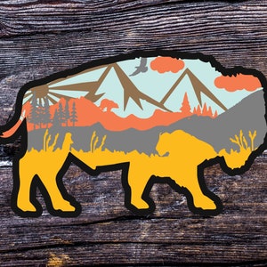 May include: A silhouette of a bison with a mountain landscape inside. The bison is yellow, the mountains are blue, grey, and brown, and the sky is light blue with white clouds.