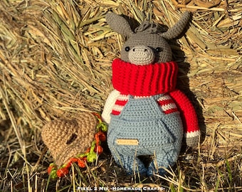 May include: A gray crocheted donkey wearing blue overalls, a red and white striped sweater, and a red scarf. The donkey is sitting in a pile of hay.