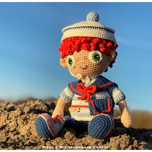 Op de afbeelding: Een gehaakte pop met rood haar, een witte matrozenpet en een blauw-wit gestreept matrozenoverhemd. De pop zit op een zanderige ondergrond.