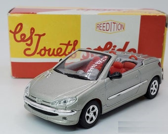 May include: A silver toy car model of a Peugeot 206 CC convertible with red interior. The car is on a white surface and the box it came in is visible in the background. The box is yellow with red accents and the text "Les Jouets" and "Reedition" is visible.