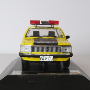 May include: Yellow and black Ford model car with a red and white police light on top. The car has a black stripe across the hood and the words "RODOVIARIA" on the stripe. The car has a license plate that reads "BA-SALVADOR GA 1983".