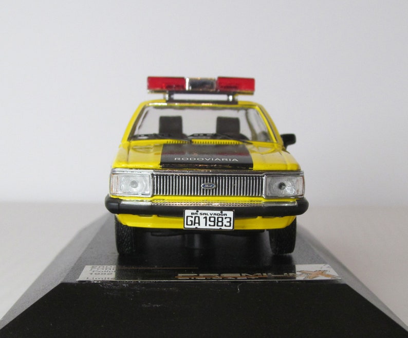 May include: Yellow and black Ford model car with a red and white police light on top. The car has a black stripe across the hood and the words "RODOVIARIA" on the stripe. The car has a license plate that reads "BA-SALVADOR GA 1983".