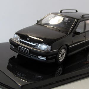 May include: A black die-cast model of an Omega estate car. The model features silver wheels, a black roof rack, and the word "OMEGA" on the front. The car is presented on a black, angled base.