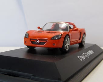 May include: A small, orange Opel Speedster model car on a black display stand. The car has a sleek design with silver wheels and a detailed interior. The stand has the text "Opel Speedster" in white.