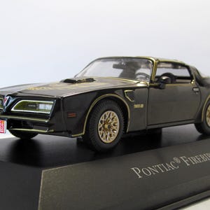 May include: A black and gold 1977 Pontiac Firebird model car on a black display stand. The model has gold-colored wheels and trim, with a license plate that reads FRI23. The car is a coupe with a sleek design.