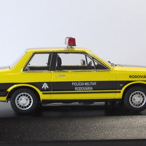May include: A yellow and black 1/18 scale diecast model car of a Ford Corcel II, a Brazilian version of the Ford Pinto. The car is a replica of a Brazilian highway patrol car, with the words "Policia Militar Rodoviaria" and "Rodoviaria" on the side. The car has a red light on top.
