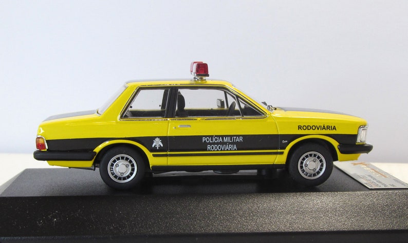May include: A yellow and black 1/18 scale diecast model car of a Ford Corcel II, a Brazilian version of the Ford Pinto. The car is a replica of a Brazilian highway patrol car, with the words "Policia Militar Rodoviaria" and "Rodoviaria" on the side. The car has a red light on top.