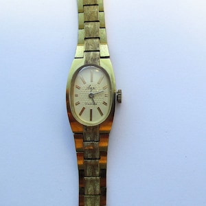 May include: A gold-toned wristwatch with a white face and black hands. The watch has a gold-toned metal band and a rectangular face. The face of the watch has a small, textured pattern and the text "Агат 17 камней" printed on it.