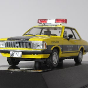 May include: A yellow and black diecast model car of a Brazilian police vehicle. The car has a red and white light bar on the roof and a license plate that reads "GA 1983". The car is labeled "RODOVIARIA" and "POLICIA RODOVIARIA FEDERAL".