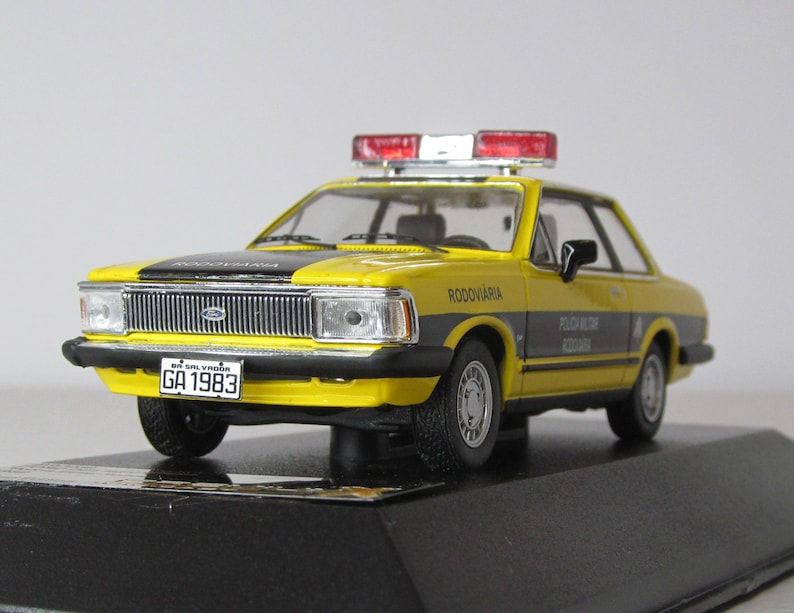 May include: A yellow and black diecast model car of a Brazilian police vehicle. The car has a red and white light bar on the roof and a license plate that reads "GA 1983". The car is labeled "RODOVIARIA" and "POLICIA RODOVIARIA FEDERAL".