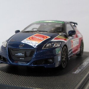以下が含まれることがあります： ENEOSやSUSUKAなどのスポンサーロゴが入った、青いホンダCR-Zレーシングカーの詳細なモデル。 ミニカーは黒いディスプレイベースに置かれています。 車は黒いホイールとリアスポイラーを備えています。