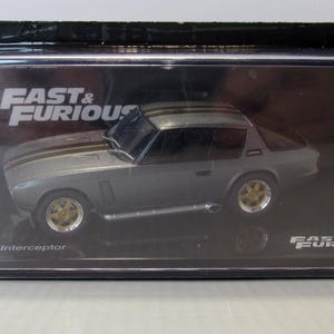Può includere: Un modellino di auto Jensen Interceptor argentato in una teca trasparente con base nera. L'auto ha interni neri e ruote dorate. La teca ha le parole "Fast & Furious" stampate sul davanti.