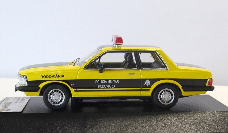 May include: Yellow and black diecast model car of a Ford Corcel, a Brazilian car, with a red light on top. The car is labeled "Pol&iacute;cia Militar Rodovi&aacute;ria" and "Rodovi&aacute;ria".