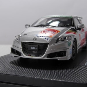 以下が含まれることがあります： 黒いテクスチャベースに展示された、レースデカール付きのシルバーのホンダCR-Zモデルカー。モデルは、詳細なヘッドライト、フロントグリル、スポンサーロゴを備えています。車はレーシングカーのミニチュアレプリカです。