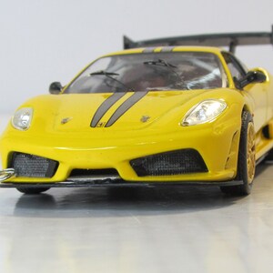 May include: A yellow toy car with black racing stripes and a rear wing. The car has a detailed front end with headlights and a grill.