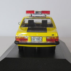 May include: Yellow toy car model of a Brazilian police car with a black stripe and the word "rodoviaria" on the back. The car has a red and blue light bar on the roof and a license plate that reads "BA-SALVADOR GA 1983".