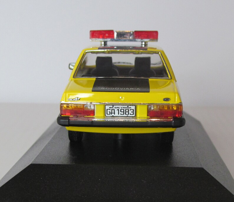 May include: Yellow toy car model of a Brazilian police car with a black stripe and the word "rodoviaria" on the back. The car has a red and blue light bar on the roof and a license plate that reads "BA-SALVADOR GA 1983".