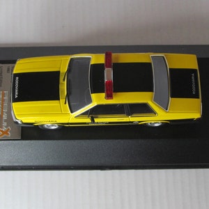 May include: A yellow and black police car model with a red light on top. The car has "RODOVIARIA" written on the side. The model is displayed in a black display case.