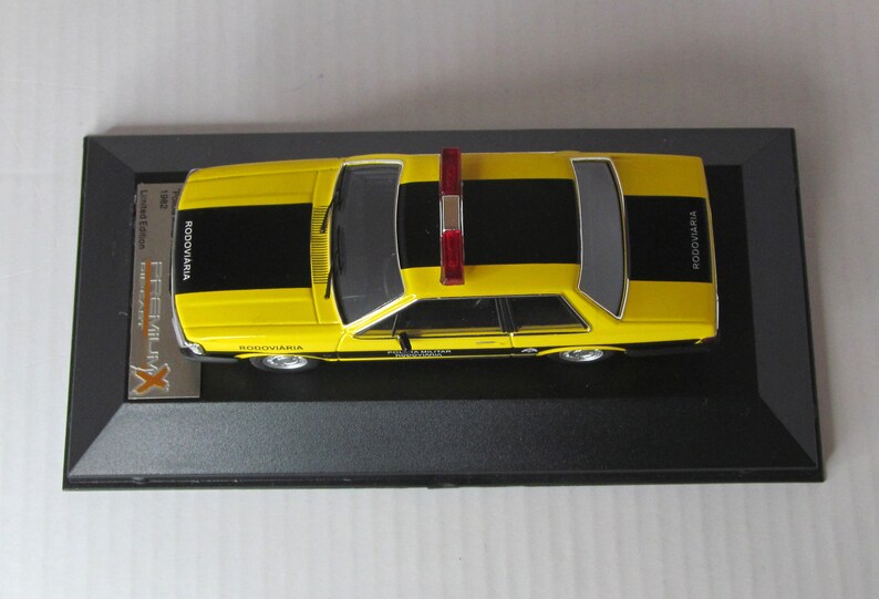May include: A yellow and black police car model with a red light on top. The car has "RODOVIARIA" written on the side. The model is displayed in a black display case.