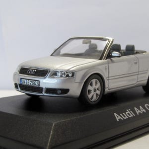 Puede incluir: Un modelo de coche Audi A4 Cabriolet plateado con interior y ruedas negros. El coche en miniatura tiene una matrícula que dice IN B2002. El modelo se exhibe sobre una base negra.