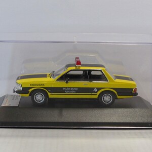 May include: A yellow and black 1/18 scale diecast model car of a Brazilian police car. The car is a Ford Corcel II, a model popular in Brazil in the 1970s and 1980s. The car has a red light on top and the words "POLICIA MILITAR RODOVIARIA" on the side.