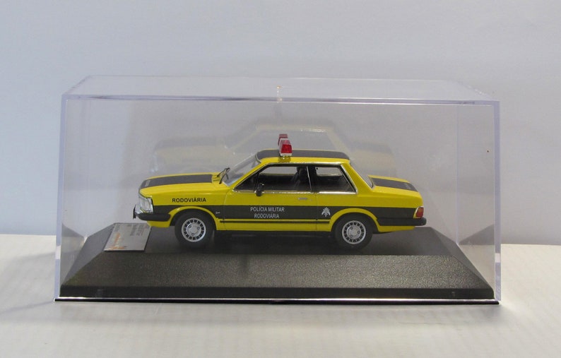 May include: A yellow and black 1/18 scale diecast model car of a Brazilian police car. The car is a Ford Corcel II, a model popular in Brazil in the 1970s and 1980s. The car has a red light on top and the words "POLICIA MILITAR RODOVIARIA" on the side.
