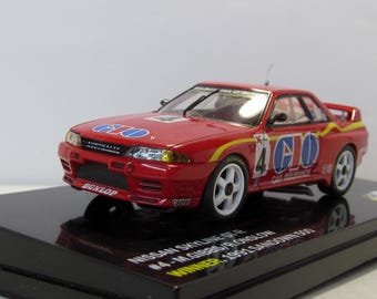 May include: A red 1/43 scale diecast model car of a Nissan Skyline GT-R race car. The car has a yellow stripe and blue and white "C.L.O." logos on the side. The car is on a black display base with the text "#4. M. GIBBS/R. PHELAN WINNER 1991 SANDOWN 500".