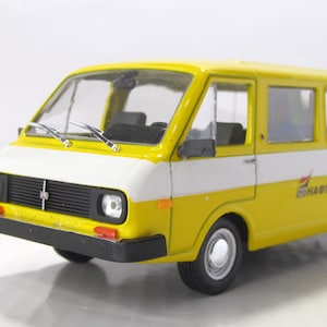 May include: A yellow and white scale model van featuring a black front grille, black bumpers, and silver-colored wheels. The side of the vehicle displays the text NAFTA in black lettering with a small red graphic element above the letters.