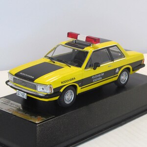 May include: A yellow and black 1/18 scale diecast model car of a Ford Corcel II, a Brazilian police car. The car has a black and white "Policia Militar Rodoviaria" sticker on the side and a red and white light bar on the roof. The car is on a black display base.