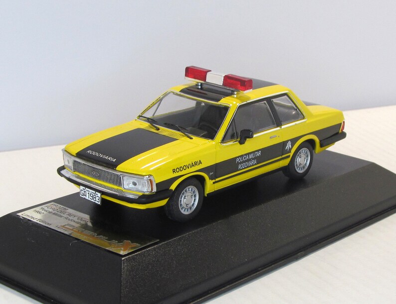 May include: A yellow and black 1/18 scale diecast model car of a Ford Corcel II, a Brazilian police car. The car has a black and white "Policia Militar Rodoviaria" sticker on the side and a red and white light bar on the roof. The car is on a black display base.