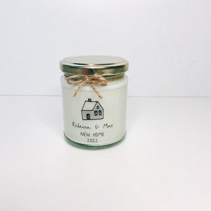 May include: A white candle jar with a gold lid and a brown twine bow. The jar has a black and white illustration of a house with the text "Rebecca & Max New Home 2022".