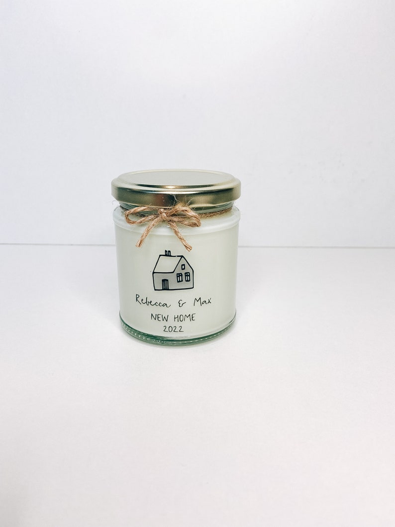 May include: A white candle jar with a gold lid and a brown twine bow. The jar has a black and white illustration of a house with the text "Rebecca & Max New Home 2022".
