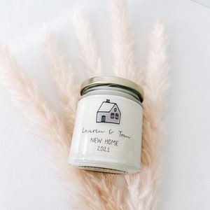 May include: A white candle jar with a gold lid. The jar has a black and white illustration of a house with the text "Lauren & Tom New Home 2021" on the front.