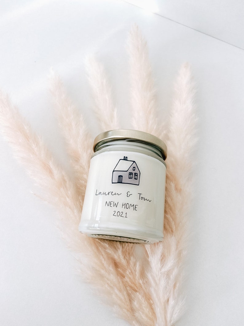 May include: A white candle jar with a gold lid. The jar has a black and white illustration of a house with the text "Lauren & Tom New Home 2021" on the front.