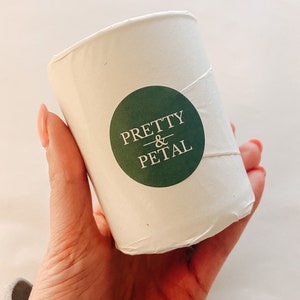 May include: A white candle container with a green circle sticker that says "PRETTY & PETAL" in white text.