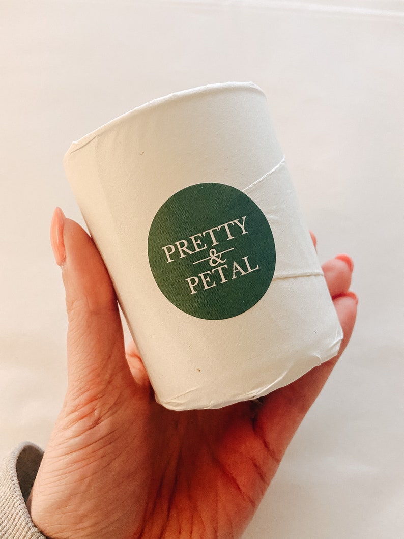 May include: A white candle container with a green circle sticker that says "PRETTY & PETAL" in white text.