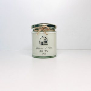 May include: A white candle in a glass jar with a gold lid. The jar has a handwritten label that says "Rebecca & Max New Home 2022" and a simple drawing of a house.