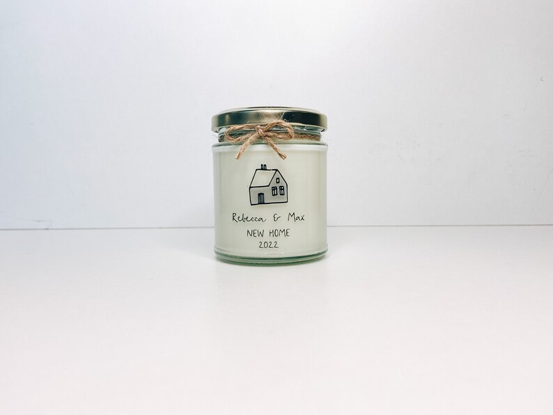 May include: A white candle in a glass jar with a gold lid. The jar has a handwritten label that says "Rebecca & Max New Home 2022" and a simple drawing of a house.