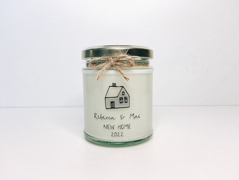 May include: A white candle in a glass jar with a gold lid. The jar is tied with twine and has a handwritten label that says "Rebecca & Max NEW HOME 2022" and a drawing of a house.