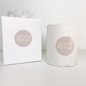 May include: A white candle with a beige label that says "Pretty & Petal" in white text. The candle is in a white box with a white ribbon.