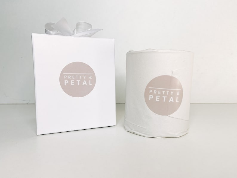 May include: A white candle with a beige label that says "Pretty & Petal" in white text. The candle is in a white box with a white ribbon.