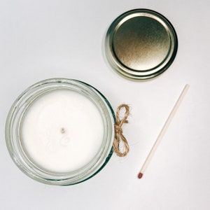 May include: A white candle in a clear glass jar with a gold lid and a brown twine bow. A match is lying on a white surface.