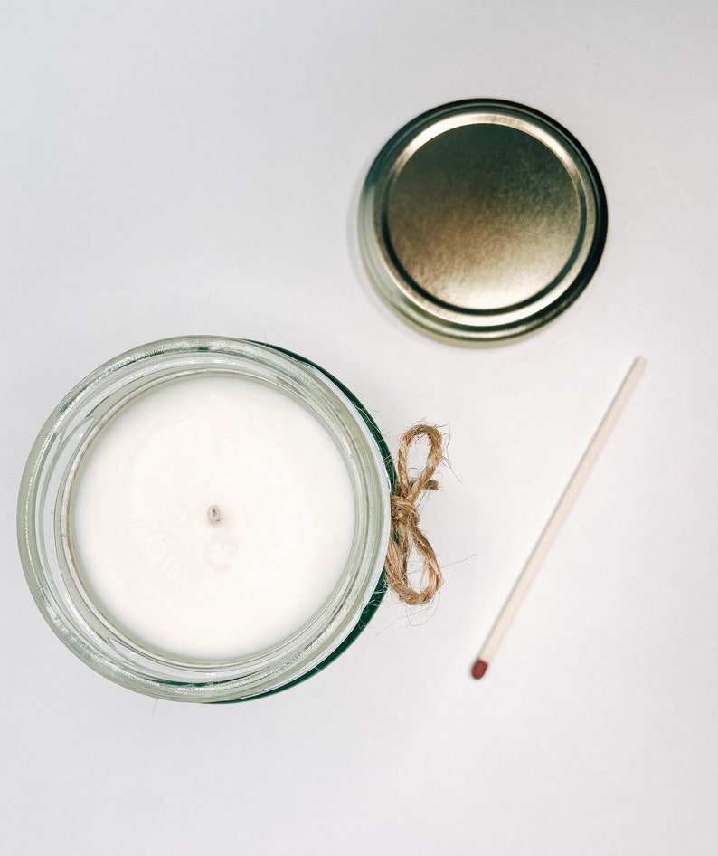 May include: A white candle in a clear glass jar with a gold lid and a brown twine bow. A match is lying on a white surface.