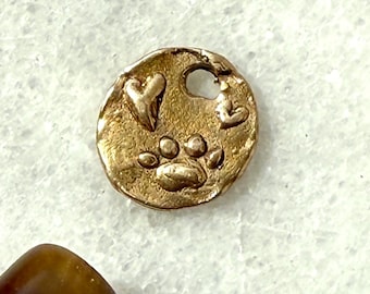 May include: A small, round, gold-colored charm with a paw print and two hearts. The charm has a small hole at the top. The charm is on a white surface.
