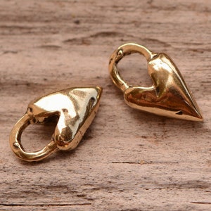 May include: Two gold-colored heart-shaped charms with a loop at the top. The charms are made of a shiny metal and are resting on a wooden surface. The heart shape is elongated with a pointed bottom.