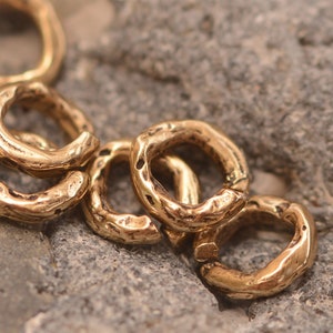 May include: Close-up of several gold-coloured, textured metal rings. The rings have an irregular, organic shape and are scattered on a grey, rough stone surface. The rings appear to be jewellery components or decorative elements.